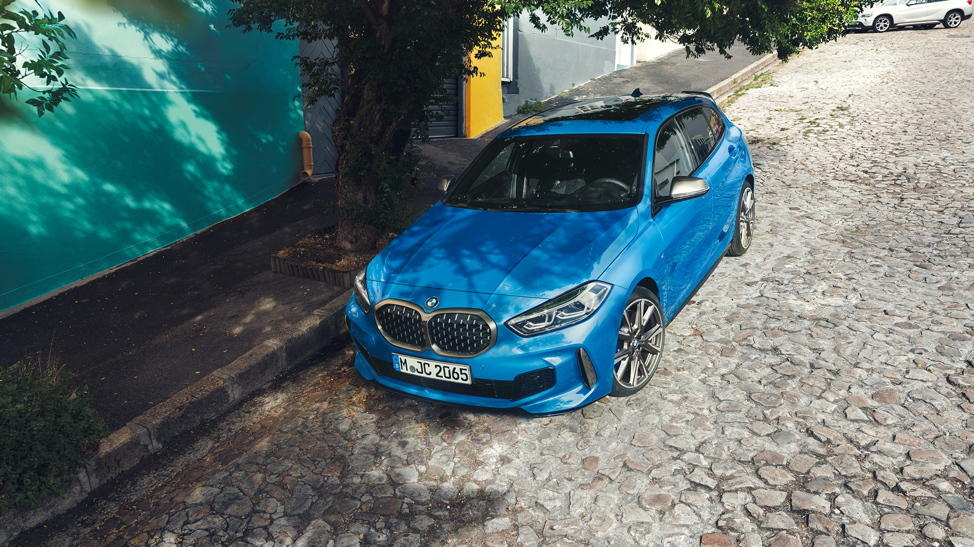 O BMW Série 1, à beira de uma rodovia. O BMW Série 1 na tonalidade Misano Blue metálico, numa vista aérea frontal inclinada, estacionado à beira de uma rodovia.
