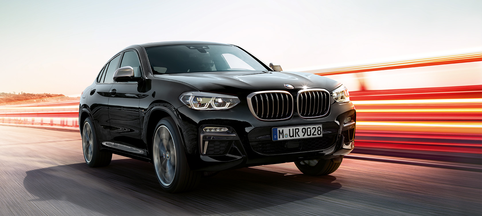 BMW X4 M40i e BMW X4 M40d, visão frontal de três quartos, lado esquerdo. BMW X4 M40i e BMW X4 M40d em preto safira metálico, exterior, visão frontal de três quartos, lado esquerdo com tiras de luz.