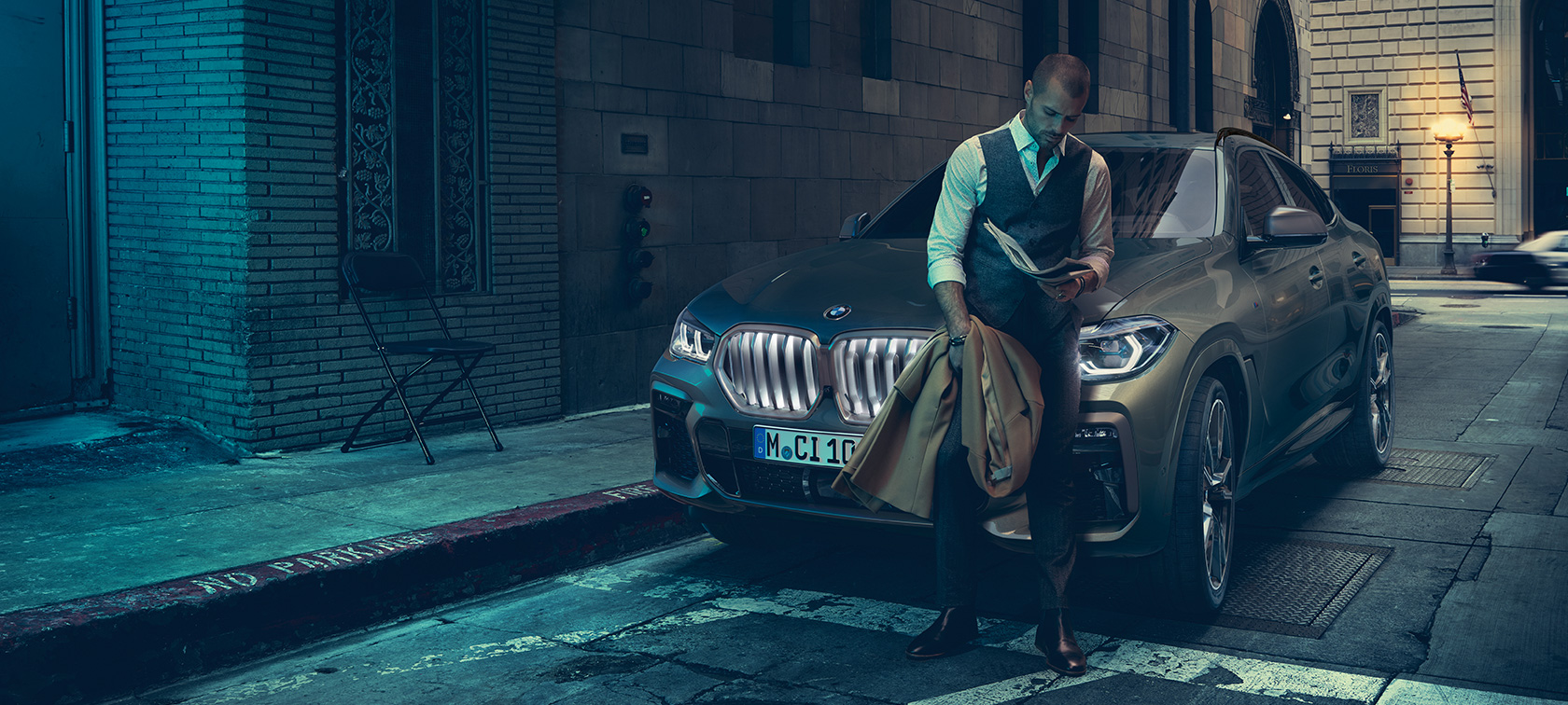 Homem encostado no BMW X6. Man leaning on BMW X6 in lateral perspective in front of an urban setting.