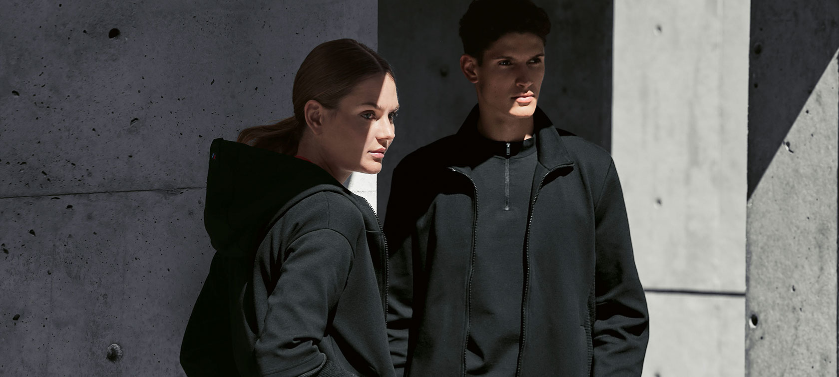Jaqueta feminina da linha BMW M e jaqueta masculina da linha BMW M. Um casal está de pé sob uma sombra. Uma mulher apresenta a jaqueta feminina BMW M e um homem veste a jaqueta masculina da linha BMW M.