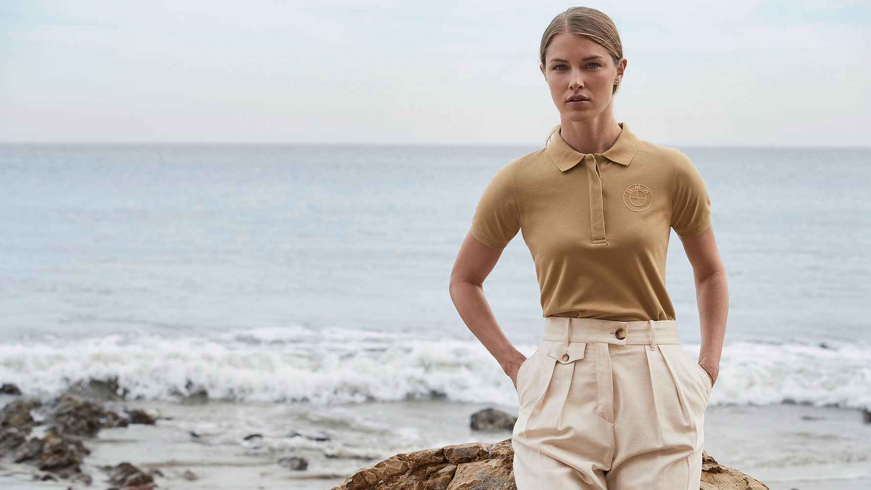 Uma mulher jovem usa o logotipo BMW em sua Camisa Polo feminina, em frente a um cenário de praia. Uma mulher jovem usa o logotipo BMW em sua Camisa Polo feminina, em frente a um cenário de praia.