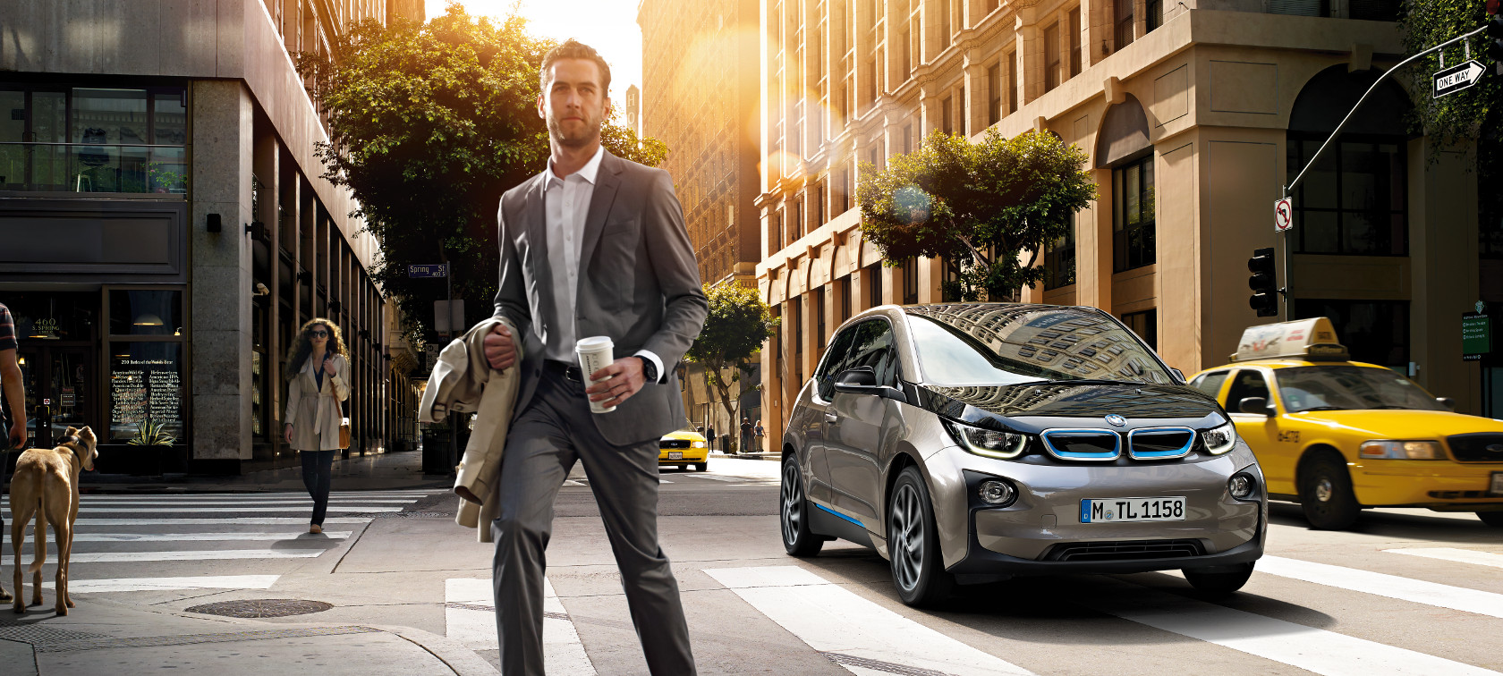 BMW i3 trafegando por cidade em meio a taxis, com homem de terno atravessando a rua, simbolizando mobilidade integrada BMW BMW i3 trafegando por cidade em meio a taxis, com homem de terno atravessando a rua, simbolizando mobilidade integrada BMW
