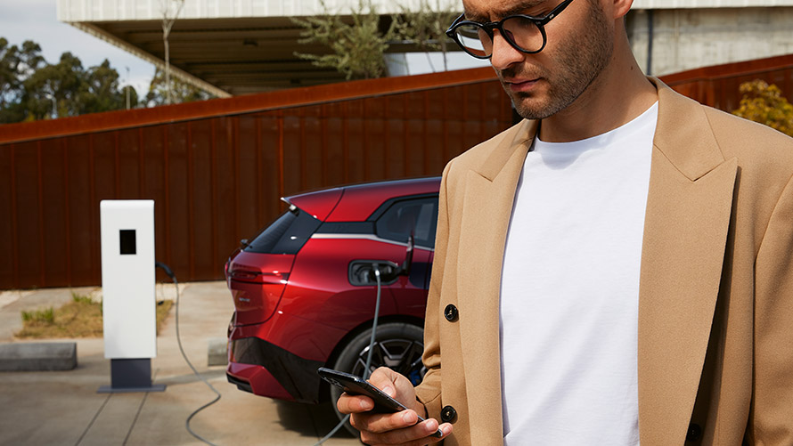 Homem com um smartphone em frente ao BMW iX, numa estação de carregamento Homem com um smartphone em frente ao BMW iX, numa estação de carregamento