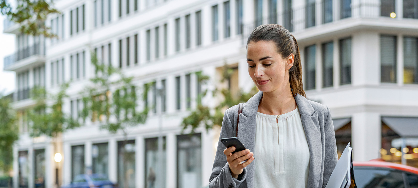 Mulher em frente a um prédio, olhando para o seu smartphone Mulher em frente a um prédio, olhando para o seu smartphone enquanto realiza a atualização remota de software