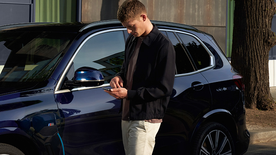 Homem apoiado na traseira do BMW e olhando seu smartphone Atualização Remota de Software Homem apoiado na traseira do BMW e olhando seu smartphone