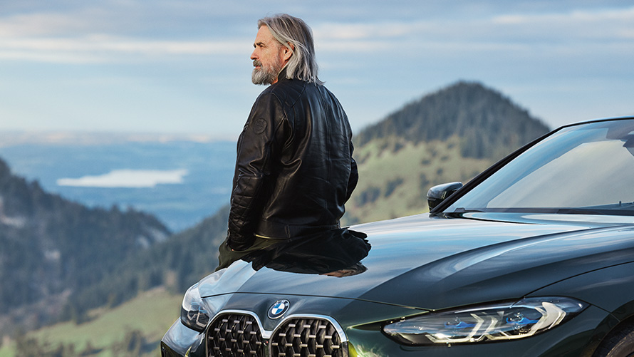 Homem inclinando-se sobre um BMW, em frente a um cenário de montanha Acessórios primavera/verão BMW, mostrando um homem inclinando-se sobre um BMW, em frente a um cenário de montanha